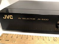 Close-up of front left side showing JVC logo and AV Selector JX-S100 model name with ruler for scale.