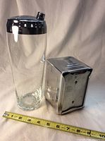 Glass cocktail shaker with metal lid next to metal napkin dispenser on white background with measuring tape showing size.