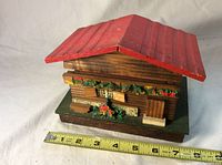 Closed Swiss chalet style music box with red roof, decorated with greenery and flowers; measuring tape included showing approx. size of 7 inches wide and 5 inches deep.
