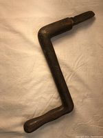 Metal hand crank viewed flat on a light cloth background showing flat profile and handle.