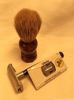 Photo shows a Gillette Super Stainless razor with white body, blade window, and a wooden-handled shaving brush with dense bristles against a beige background.