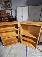 Two light wood-finished bookcases placed side by side, each containing three shelves. Visible wear and marks on shelves and outer surfaces.