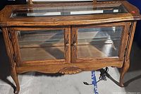 Front view of the wooden curio cabinet showing glass doors with brass handles and carved motif on front panel.