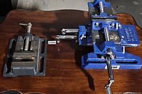 Two drill press vises on wooden table, blue Central Forge and smaller gray vise.