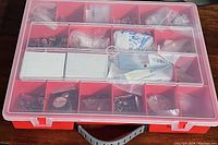 Top view of closed red plastic storage organizer box with clear lid showing multiple compartments holding various small hardware items in plastic bags and boxes