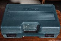 Closed dark green carrying case with embossed Hitachi logo, seen from the top, showing latches and handle.
