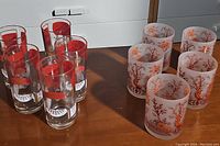 Overall view showing both vintage glass sets on a wooden surface: six frosted floral design glasses and six clear drink recipe glasses.