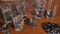 Two sets of vintage glassware displayed on wooden surface. A set of four glass mugs with intricate metallic floral holders and handles. A set of three clear drinking glasses with black leather studded holders and two separate holders.