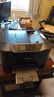 Front angled view of Canon multifunction printer on desk showing paper input and output trays, control panel, and overall compact design.