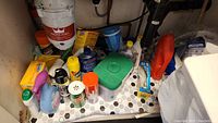 Lot of various partially used cleaning products stored under sink with visible plumbing and waste disposal unit