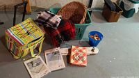 Wicker basket, plastic laundry basket, 100% wool plaid scarf, Prince of Wales wool throw blanket with holes, cooler on wheels with pull-out handle, laundry pins in blue container, mini ironing board, tri-circle lock with keys, vintage cookbook, and papers.