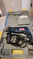 Ryobi hammer drill in open carrying case with cord and side handle visible.