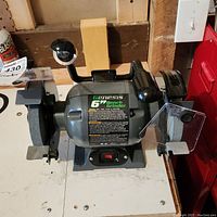 Front view of the Genesis 6.7 inch bench grinder showing both grinding wheels, safety shields, and power switch on the base.