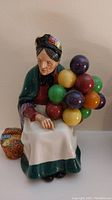 Front view showing elderly woman holding colorful balloons in multiple colors, seated with basket of flowers.