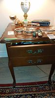 Wooden side table with drawer showing scratches, decorative items on top, partial drawer open, standing on rug.