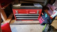 Red Craftsman metal tool box on wooden shelf with closed drawers, showing brand label and latches