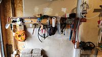 View of tools hanging on a wooden rack including garden trowels, hand rake, pruning clippers, hammer, extension cord reel, hats, and other assorted tools and accessories in a garage setting.