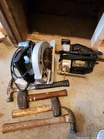 Photo showing Black & Decker circular saw, Sears Craftsman jigsaw, and four hammers with wood and rubber handles lying on a concrete floor.