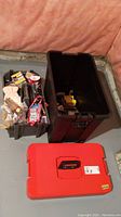 Overview of two black plastic containers including one with a red lid containing assorted hand tools and hardware pieces.