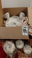 Cardboard box containing the china pieces, showing stacked plates and two teacups on top, with some packing paper