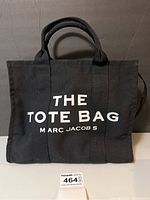 Front view of the black canvas tote bag with "THE TOTE BAG MARC JACOBS" printed in white on the front.