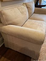 Angled front corner view showing buttercream chenille upholstery, rolled arm, loose seat and back cushions, and fringe skirt.