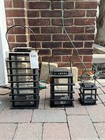 Full view of the three black metal lantern style candle holders arranged by size on a brick patio with a brick wall background.