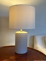Photo of the full lamp showing textured grey ceramic base and white fabric shade on round wooden table next to armchair