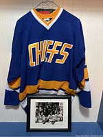 Full view of blue Charlestown Chiefs jersey on hanger with framed photo beneath