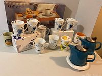 All items arranged on table: boxed mugs, loose mugs, mugs with lids and strainer, teapots, novelty mugs, folding tray behind