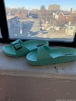 Side view of the mint green children's sandals set against a window background, showing single strap with metal buckle and contoured footbed.