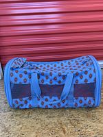 Soft-sided blue pet carrier with patterned diamond print, mesh ventilation panels, and handles lying on wooden floor against orange storage door.