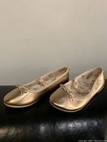 Pair of gold metallic ballet flats for girls shown side-by-side on a black surface against a plain background.