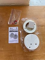 Photo shows a white circular Kidde smoke alarm, its mounting base, a small plastic bag with screws and wire connectors, and a user manual.