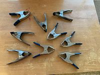 Overview of 9 metal triangle holder clamps with black and blue plastic-coated handles on a wooden surface