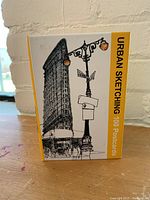 Front of the Urban Sketching 100 Postcards box showing a detailed sketch of a building and street lamp.