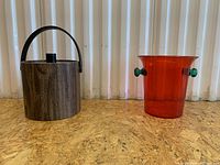 Two vintage ice buckets shown side by side on a wood surface against a light background. Left is brown woodgrain bucket with handle and lid, right is orange acrylic bucket with green side knobs.