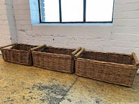 Three rectangular wicker baskets lined up on a wooden floor in front of a window with brick wall background, showing their shape, size, and wooden handles.