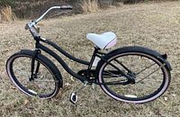 Side view of black Huffy Cranbrook women's cruiser bike with white and pink seat and pink tires in grassy outdoor setting.