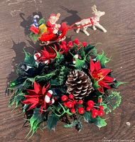 Photo showing vintage plastic candle wreath with red poinsettias, pinecones, red berries, candy canes, and holly leaves with a small Santa sleigh and reindeer figurine in the background on wood surface.