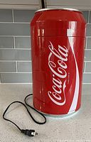 Closed Coca-Cola branded mini fridge shaped like a red soda can with power cord visible.