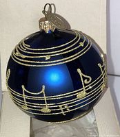 Close-up photo showing the dark blue spherical Christmas ornament decorated with glittery gold music notes and staff lines, with the Pier 1 metal tag visible on the gold ornament cap.