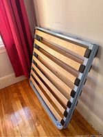 Foldable metal bed frame with wooden slats standing upright against a wall beside a window with red curtains, showing the side profile and slats partly visible.