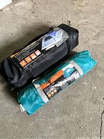 Photo showing two packed camping tents in storage bags: one black and one teal, placed side by side on a concrete floor.
