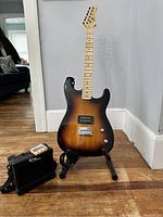 Sunburst Ryse by Sawtooth electric guitar on stand with cable and mini amplifier on wooden floor against light gray wall.