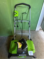 Front view of Greenworks electric snow blower showing handlebar, power cord, and front intake area with Greenworks branding.