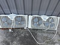 Two white window fans side by side on the ground showing front grilles and cords