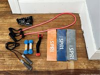 Photo showing three SPRI resistance bands, FILA fingerless workout gloves, two blue 2lb dumbbells, a red-handled jump rope, and pink resistance band with handles arranged on wooden floor.