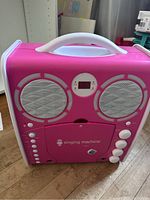 Front view of pink karaoke machine showing the speaker grills, control buttons, LCD screen, and white handle.