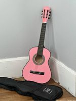 Small pink kids acoustic guitar leaning against a wall on a wooden floor, black soft carrying bag lying in front.